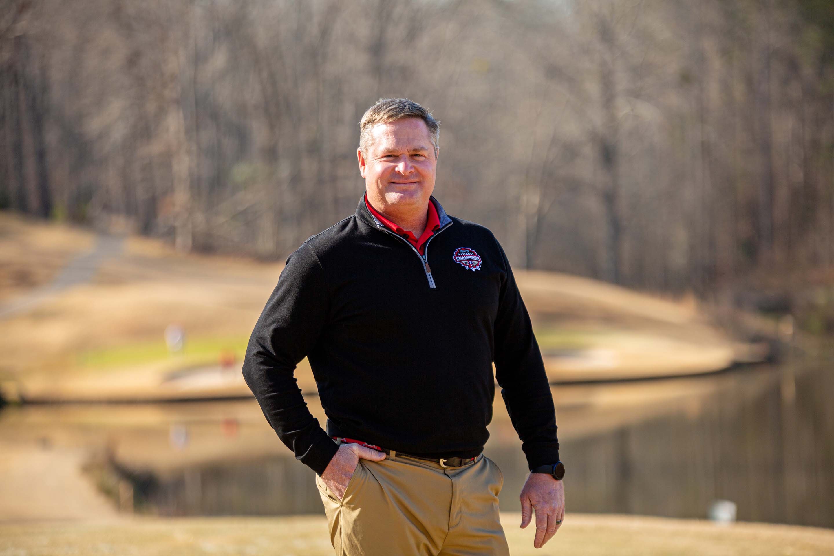 A photograph of Scott Griffith wearing a black jacket with a UGA National Champions logo patch. Behind him is a pond and a portion of the golf course green.
