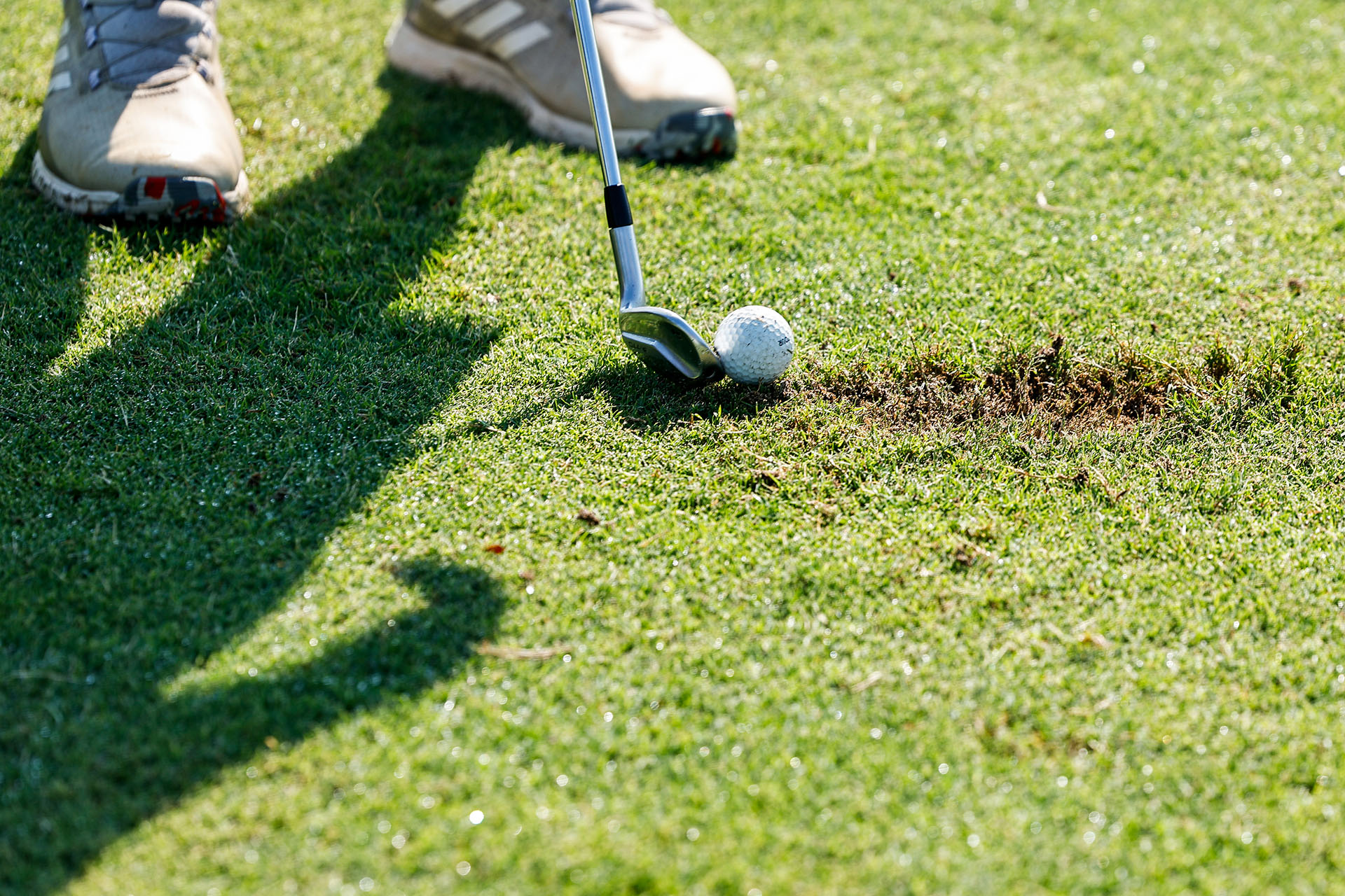 A close-up photograph of someone lining up a golf club with a golf ball on the grass. Grey shoes can be seen towards the top of the photo, and the grass to the right of the ball is scuffed.