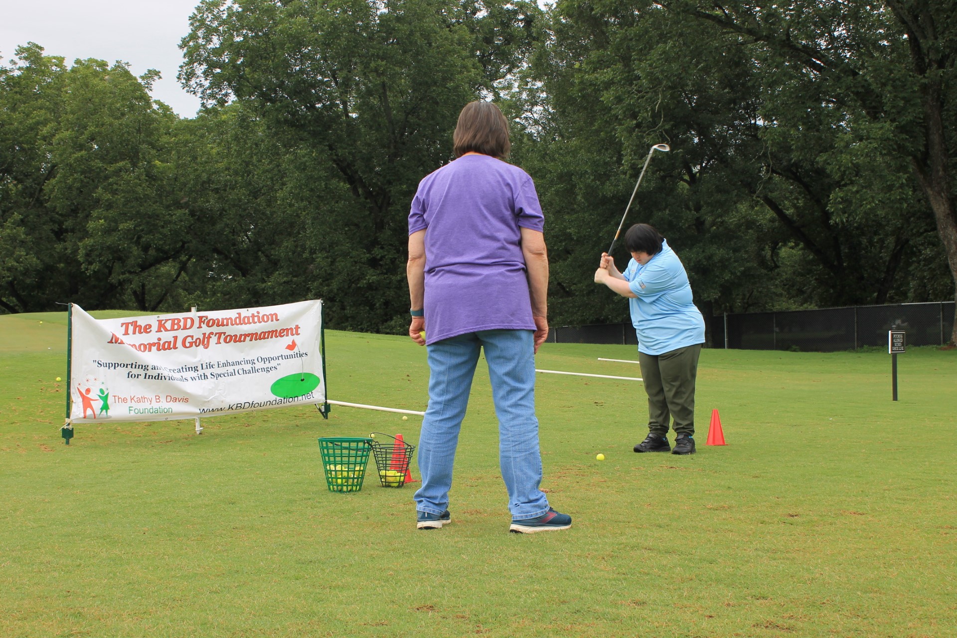 A photograph of two individuals on the putting green. One has their back to the camera and is watching the other prepare to strike a golf ball. A banner to their left reads, " The KBD Foundation Memorial Golf Tournament, 'Supporting and creating Life Enhancing Opportunities for Individuals with Special Challenges;" The Kathy B. Davis Foundation; www.KBDfoundation.net"