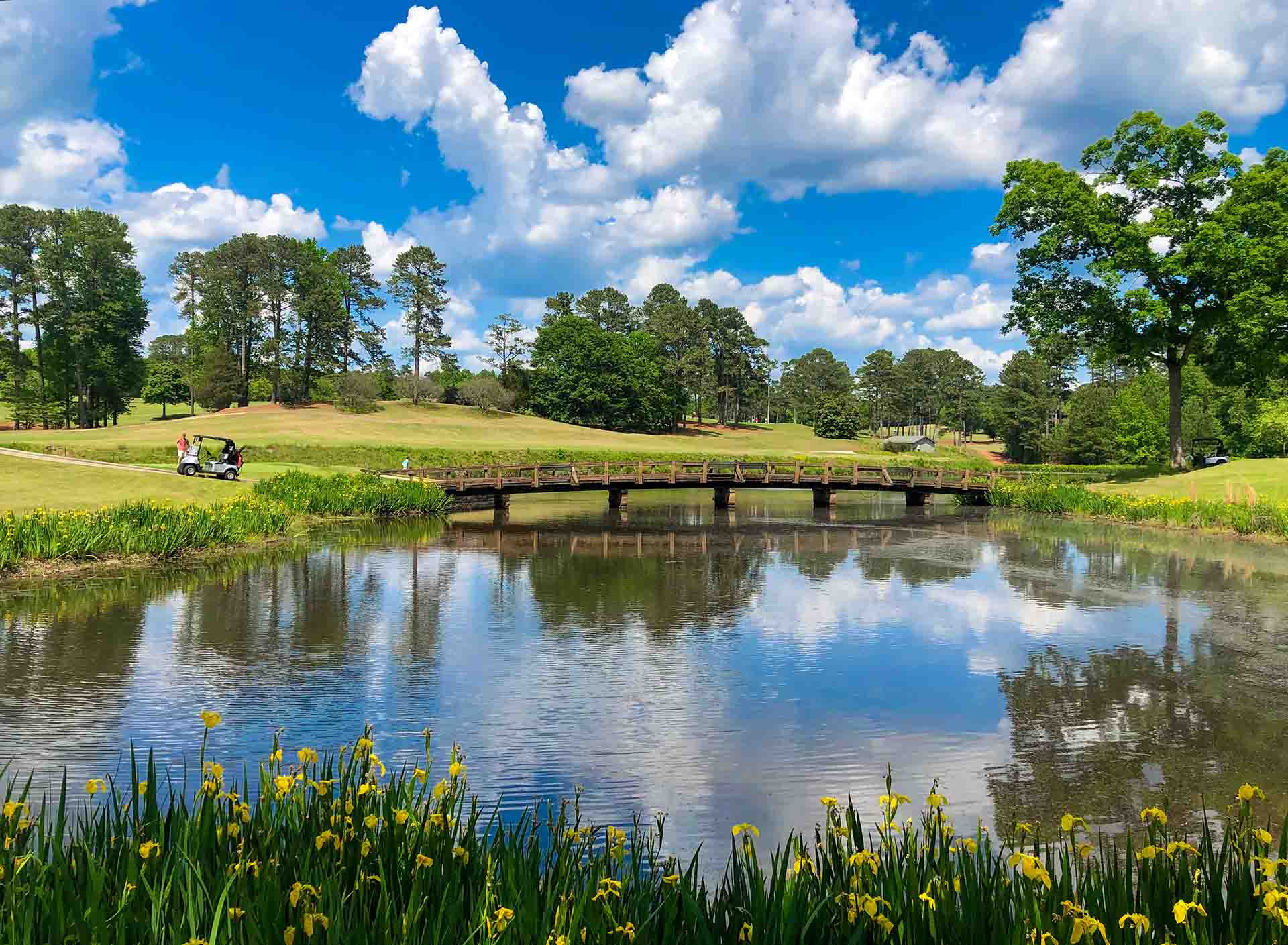 a landscape photo of the UGA golf course featuring a lake in the foreground, a wooden bridge crossing the lake, and the golf green and trees in the background. A person stands next to a golf cart on the left-hand side of the green.